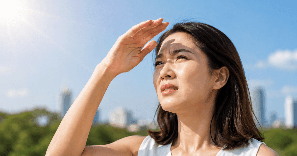 Asian woman shielding her eyes from strong sunlight outdoors.