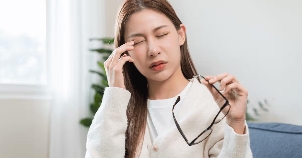 Asian woman experiencing eye pain while holding her glasses.